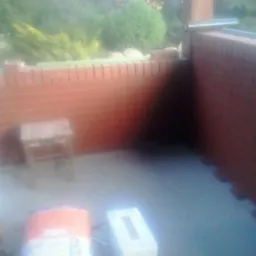 Ceglana ściana w trakcie budowy, widoczne worki z zaprawą i stołek. Budowa z czerwonej cegły, etap murowania, zieleń w tle.