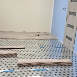 Układanie paneli podłogowych na folii izolacyjnej z widocznymi fragmentami nieułożonych paneli i niebieską taśmą mierniczą w jasnym pomieszczeniu z kremową i błękitną ścianą.