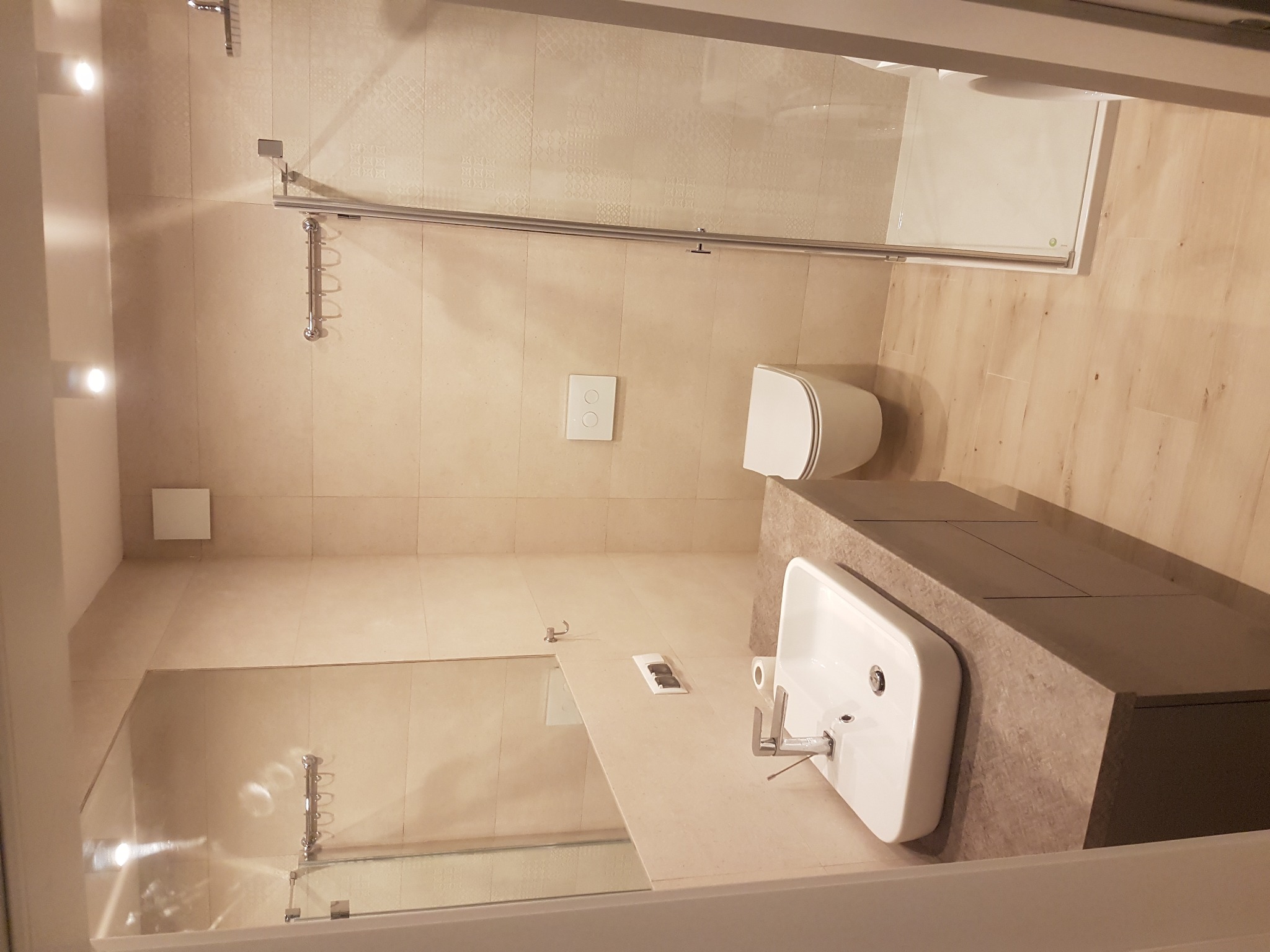 Nowoczesna łazienka z beżowymi płytkami, kabina prysznicowa ze szklanymi drzwiami, umywalka nablatowa na szarym blacie, wisząca toaleta i drewnopodobna podłoga.