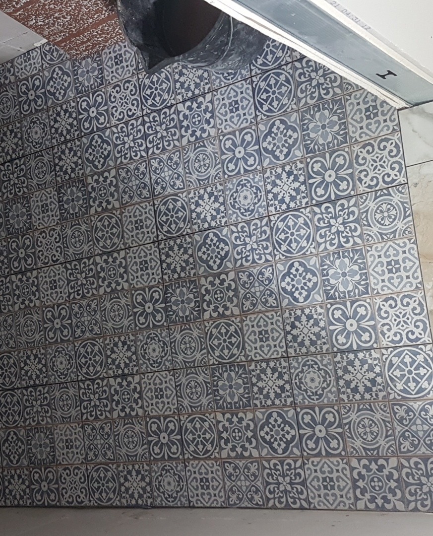 Ułożone, wzorzyste, niebiesko-białe płytki podłogowe, widoczny fragment sufitu z instalacją oraz wiadro budowlane w rogu.