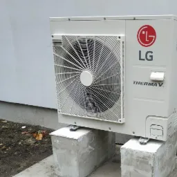 Zewnętrzna jednostka pompy ciepła LG Therma V, zamontowana na betonowych bloczkach przy szarej ścianie budynku. Widoczny wentylator i logo producenta.