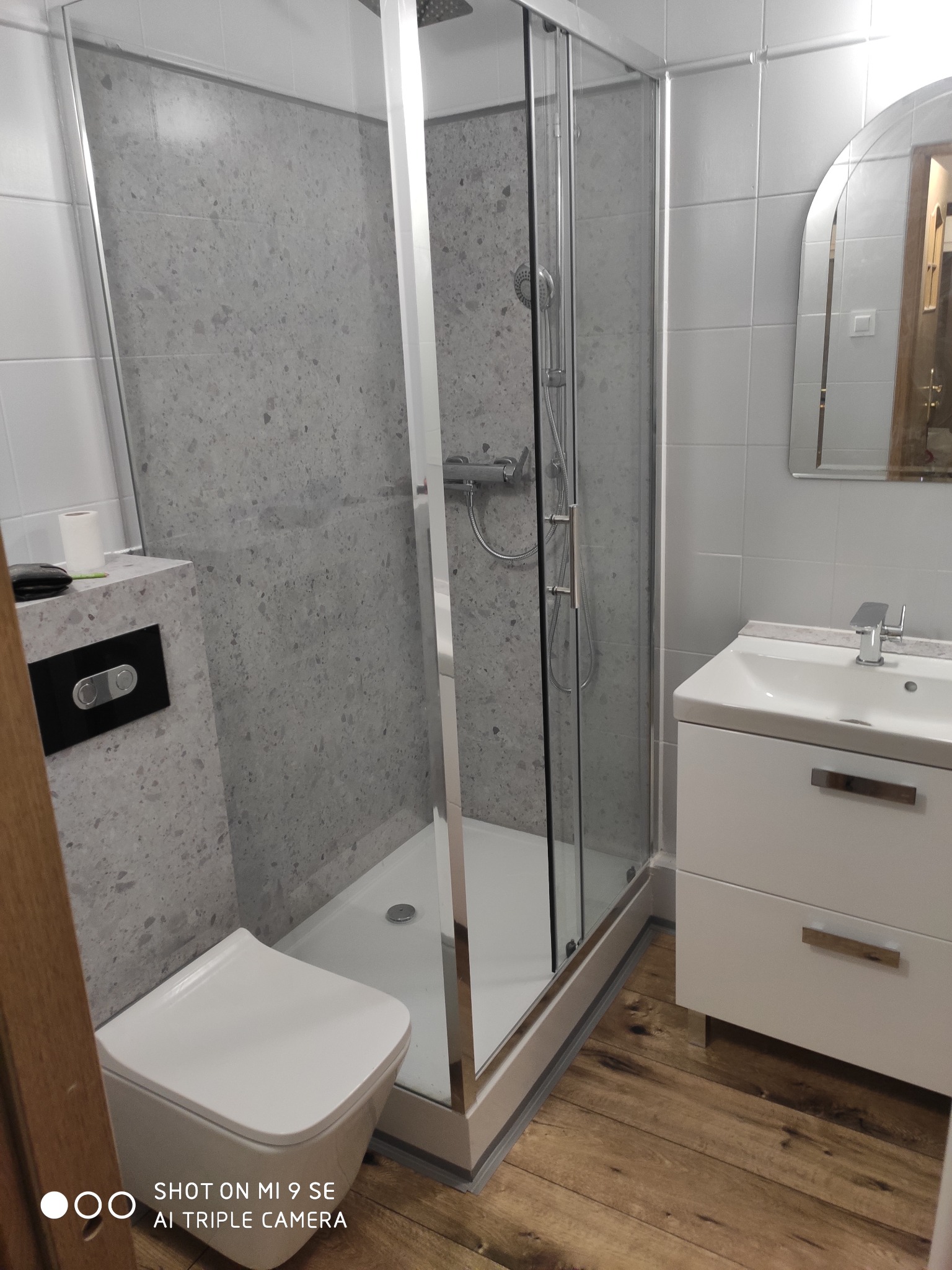 Nowoczesna łazienka po remoncie: kabina prysznicowa ze szklanymi drzwiami, umywalka z szafką, toaleta podwieszana i drewnopodobna podłoga. Szare płytki na ścianach.