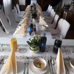 Długi stół nakryty białym obrusem z dekoracyjnym bieżnikiem, zastawiony naczyniami z zupą, napojami w szklanych karafkach i butelkach, kwiatami w doniczkach, przykryty krzesła białymi pokrowcami...