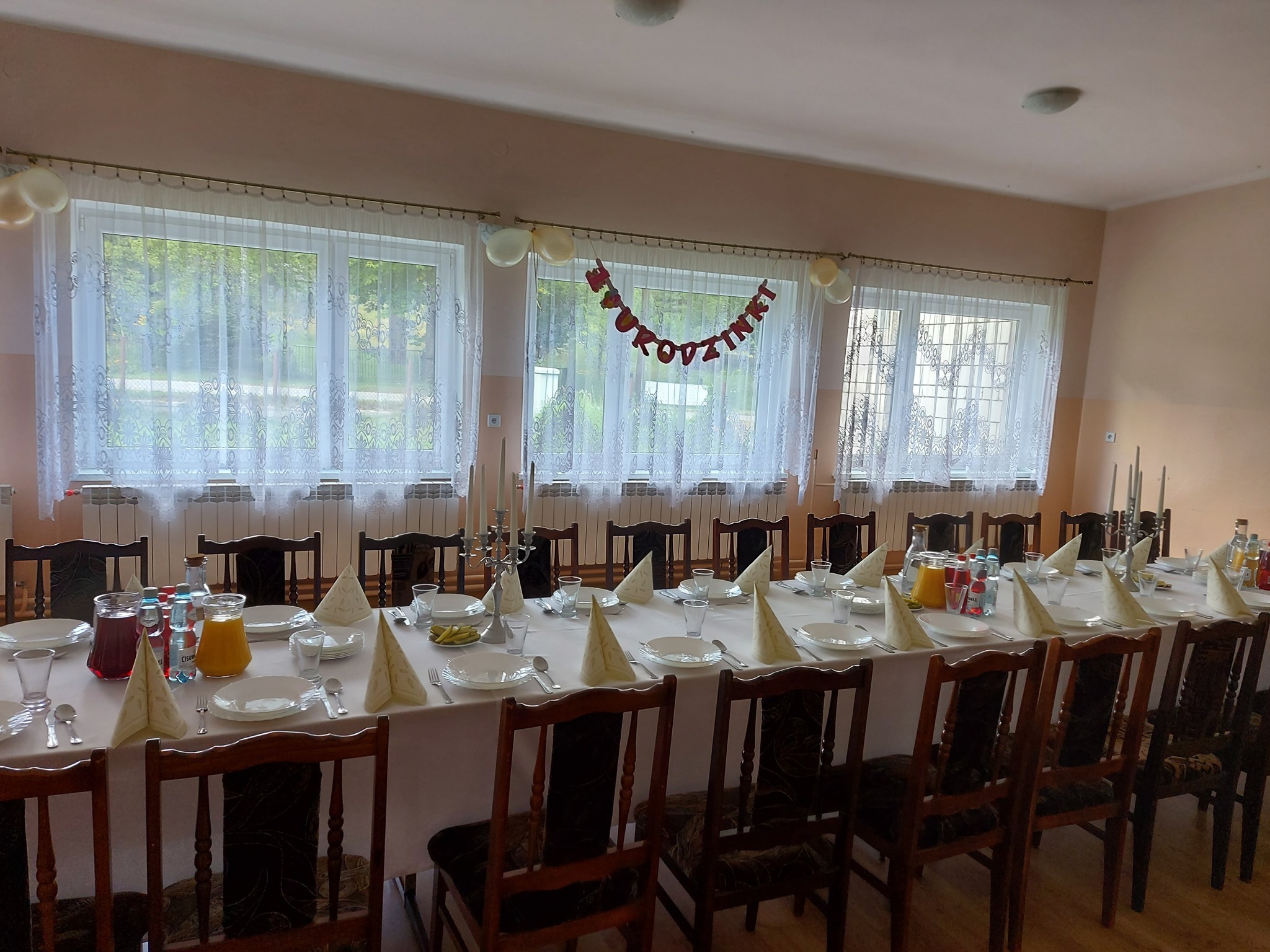 Długi stół nakryty białym obrusem, ustawiony w sali z oknami, przygotowany na uroczystość. Na stole talerze, sztućce, szklanki, napoje w dzbankach i butelkach, serwetki złożone w stożki. Nad oknami...