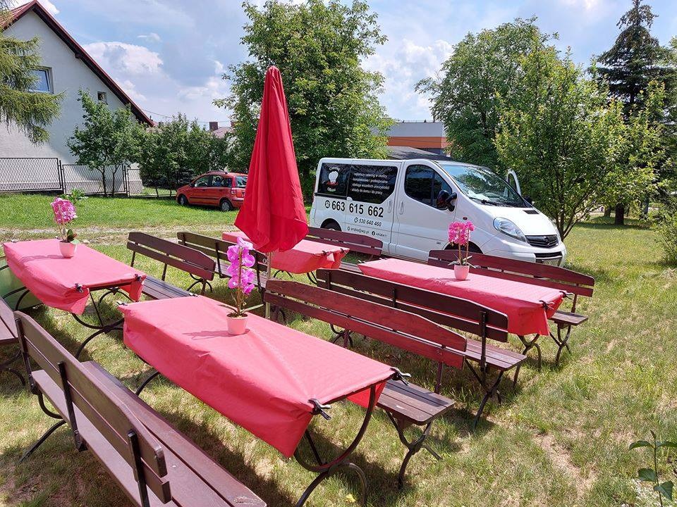 Przygotowane stoły z różowymi obrusami i kwiatami w doniczkach na zewnątrz, obok stoi biały van z numerem telefonu, czerwony parasol i budynek w tle, sugerujące catering plenerowy.