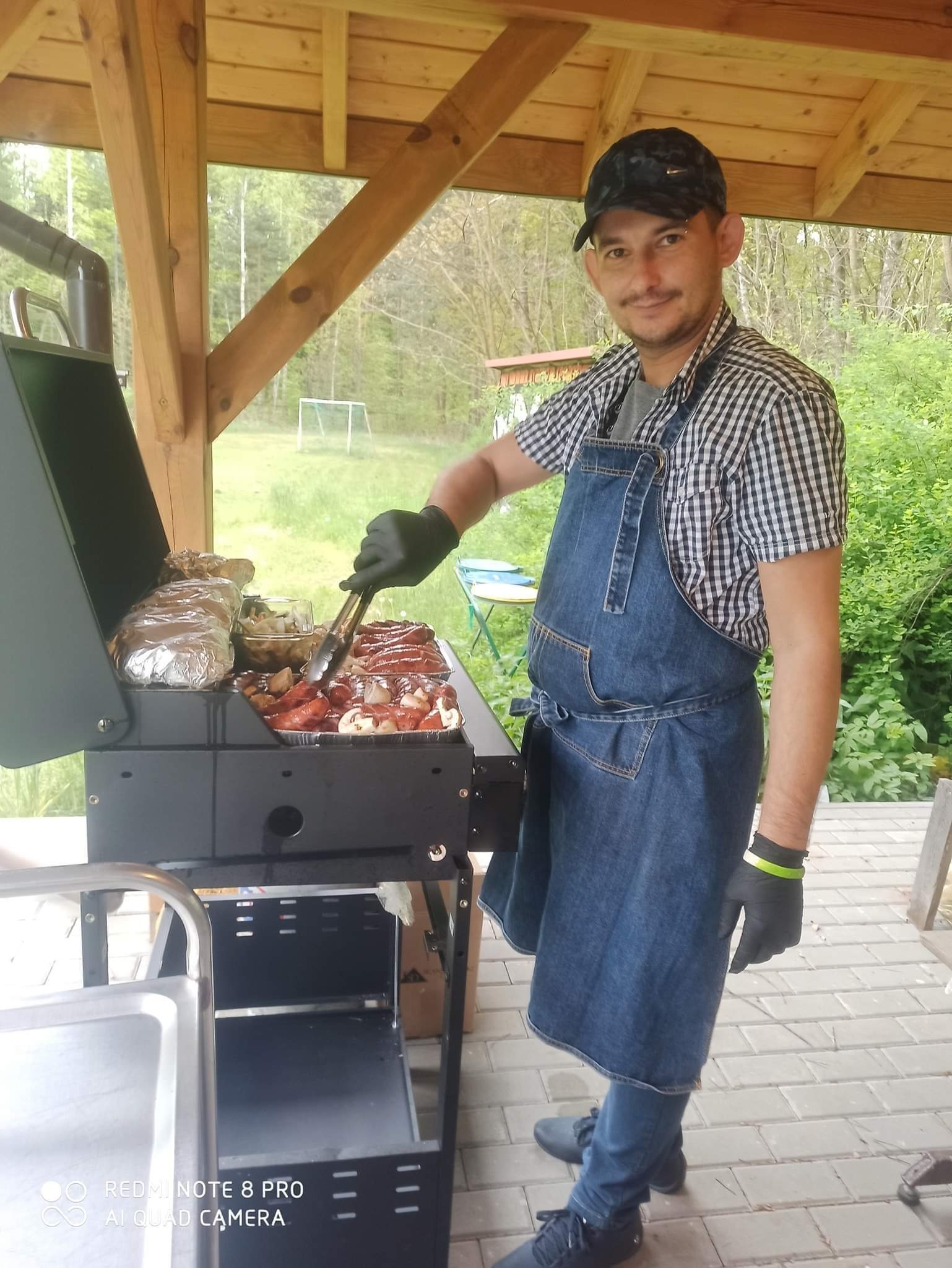 Mężczyzna w dżinsowym fartuchu i czarnych rękawiczkach grilluje kiełbaski i warzywa na czarnym grillu w drewnianej altanie, w tle zielona trawa i drzewa.
