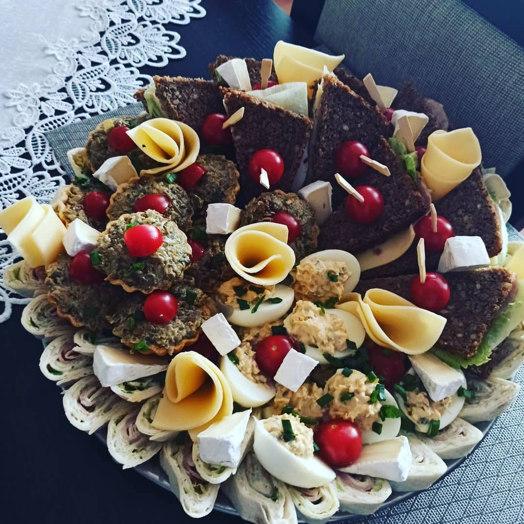 Dekoracyjna taca z przekąskami: tartaletki z pastą, kanapki z ciemnego pieczywa, roladki z tortilli, jajka faszerowane, ser brie i pomidorki koktajlowe, ułożone w formie kompozycji.