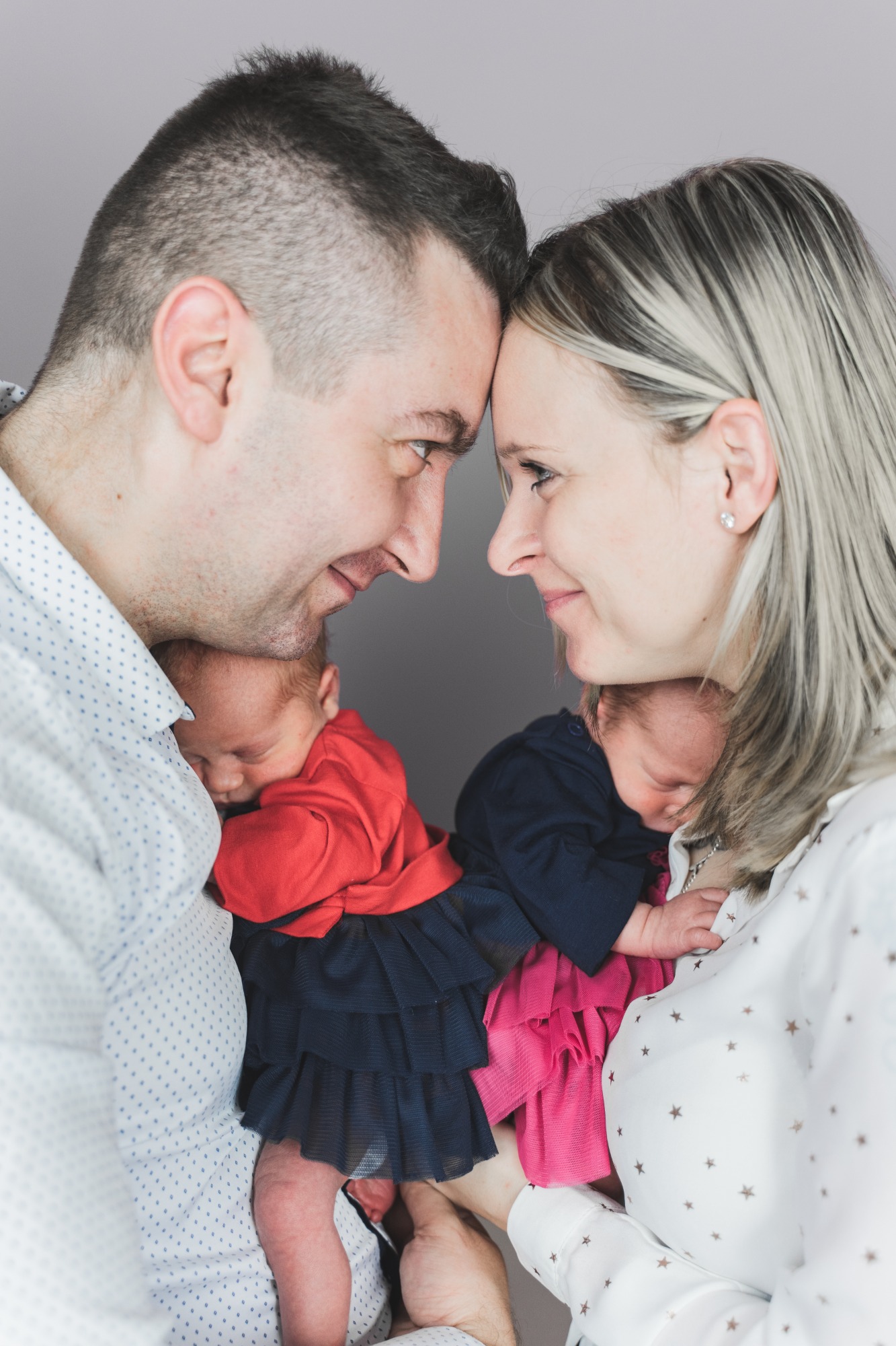 Rodzice patrzą na siebie czule, trzymając na rękach śpiące noworodki w kolorowych ubrankach na szarym tle.