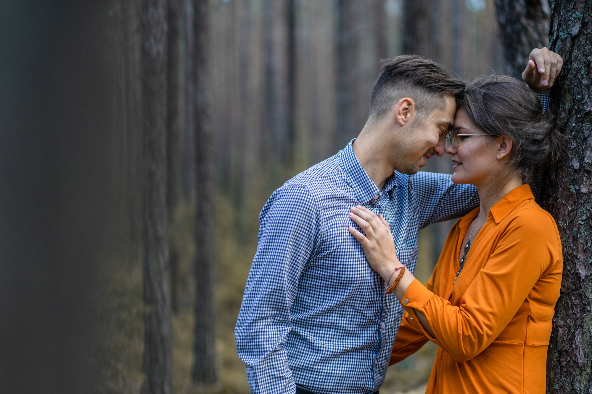 Zakochana para oparta o drzewo w lesie, kobieta w pomarańczowej sukience i okularach, mężczyzna w niebieskiej koszuli w kratę, zbliżone twarze, spokojna atmosfera.