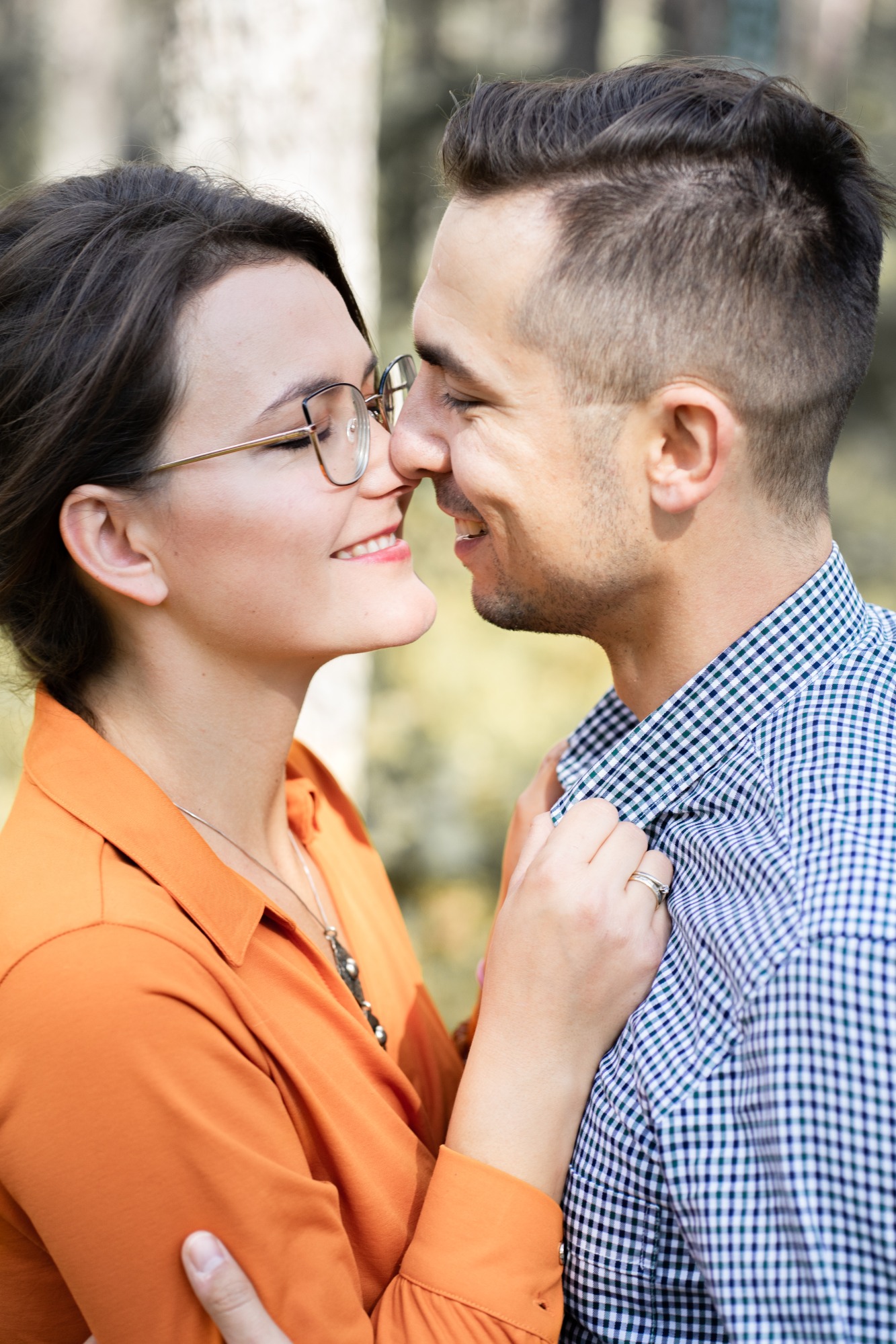 Zbliżenie na parę dotykającą się nosami; kobieta w okularach i pomarańczowej bluzce, mężczyzna w koszuli w kratę, oboje uśmiechnięci na tle drzew.