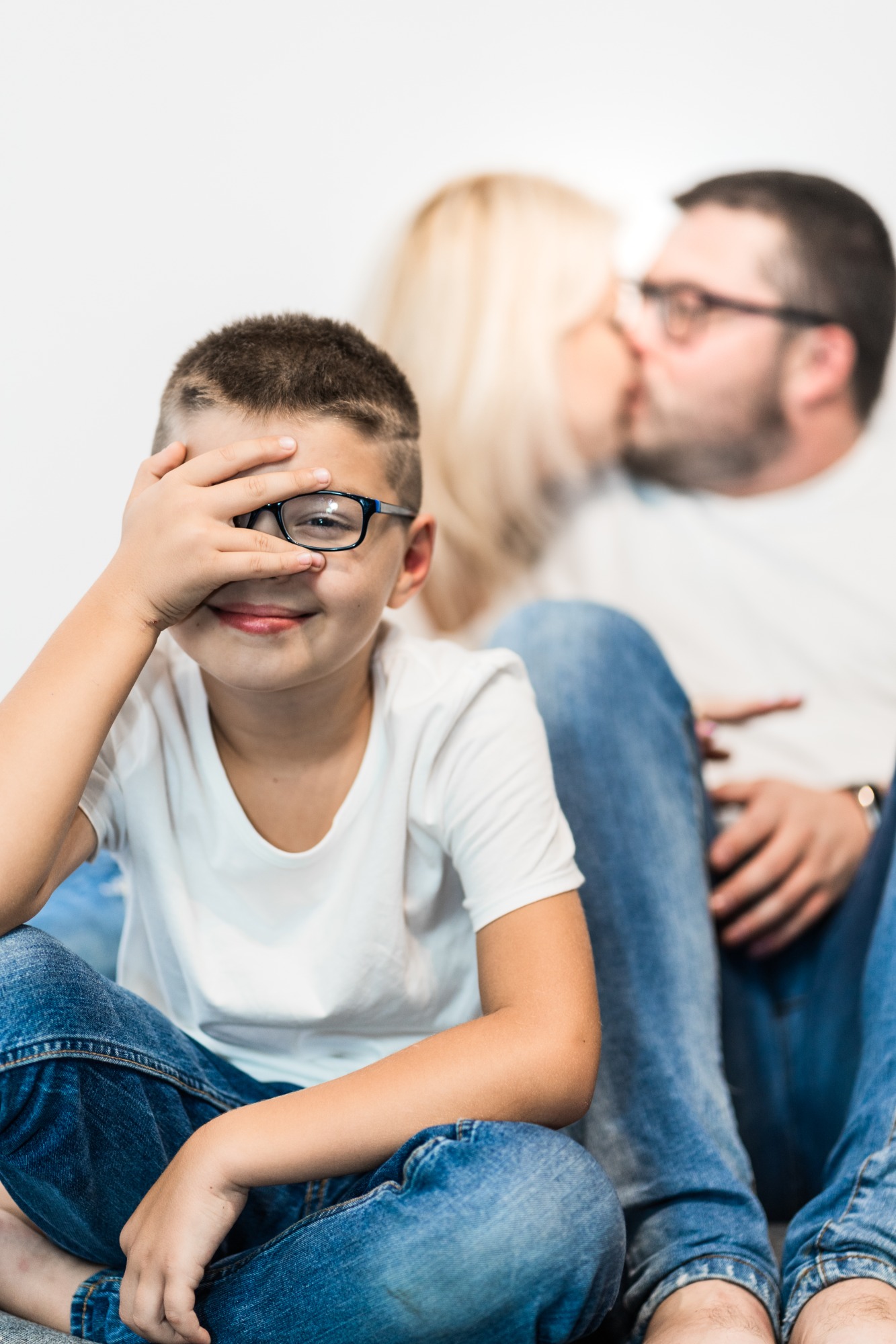 Uśmiechnięty chłopiec w okularach zakrywa jedno oko dłonią, w tle całująca się para, wszyscy ubrani w białe koszulki i dżinsy.