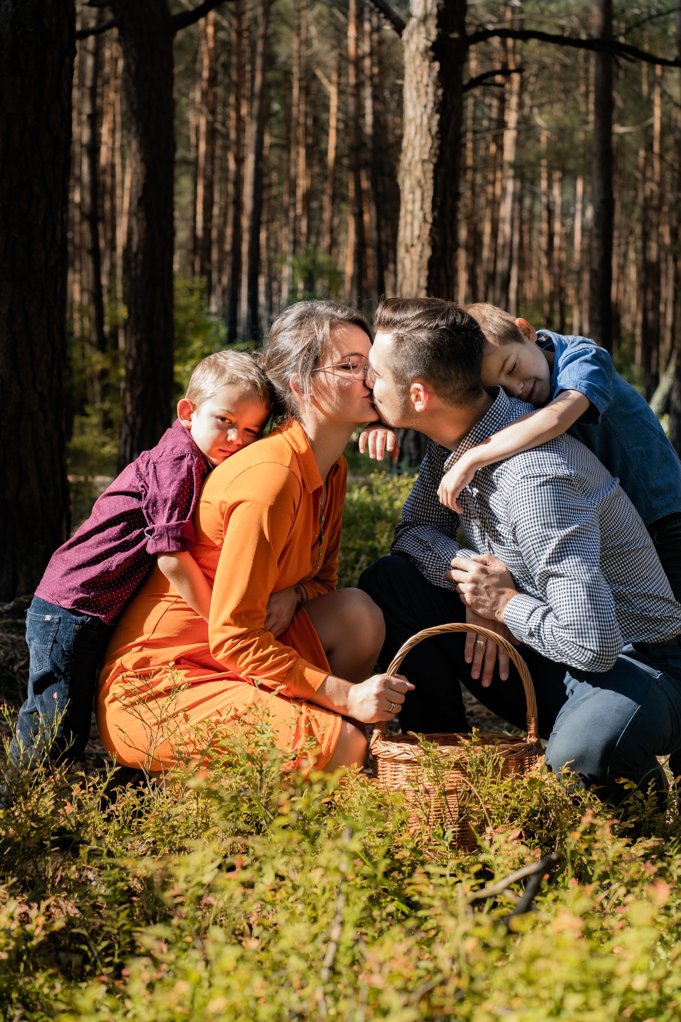 Rodzina w lesie: rodzice całują się siedząc na ziemi, przytuleni przez dwóch synów, koszyk piknikowy w pobliżu.