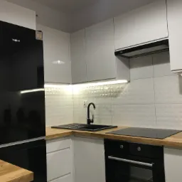 Nowoczesna, minimalistyczna kuchnia z białymi szafkami, czarnym zlewem i armaturą, drewnianym blatem oraz czarną lodówką i piekarnikiem, oświetlona listwami LED pod szafkami wiszącymi.