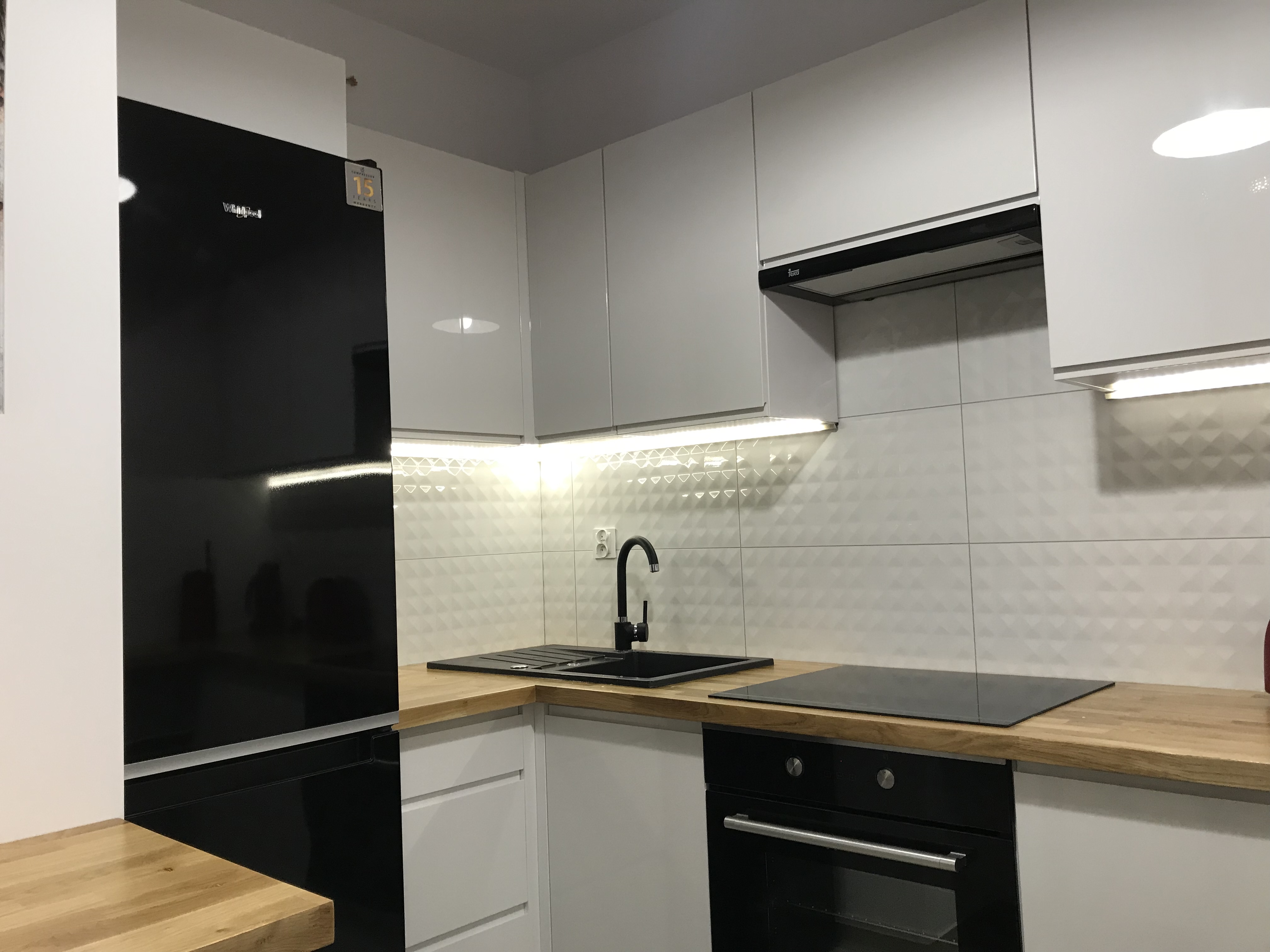 Nowoczesna, minimalistyczna kuchnia z białymi szafkami, czarnym zlewem i armaturą, drewnianym blatem oraz czarną lodówką i piekarnikiem, oświetlona listwami LED pod szafkami wiszącymi.