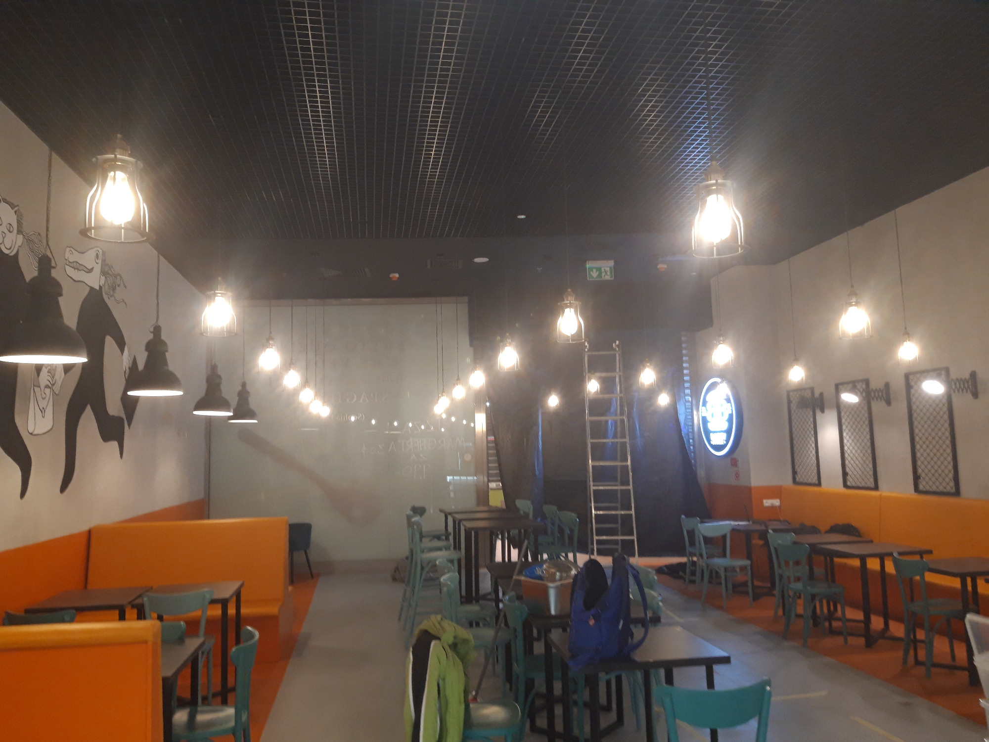 Wnętrze lokalu Bobby Burger Avenida w trakcie prac wykończeniowych: widoczne stoły, krzesła, oświetlenie, grafika na ścianie oraz drabina oparta o ścianę, sugerująca trwające prace instalacyjne...