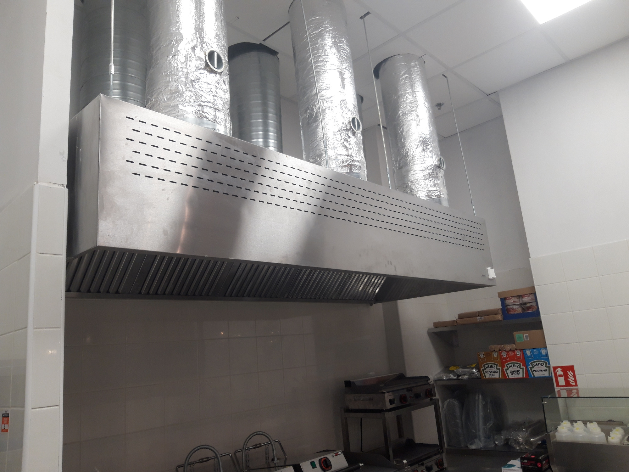 Profesjonalny system wentylacyjny ze stali nierdzewnej w kuchni komercyjnej, z widocznymi kanałami wentylacyjnymi owiniętymi folią aluminiową, zainstalowany nad sprzętem kuchennym.