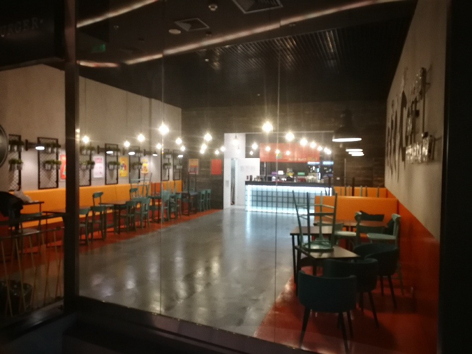 Wnętrze lokalu Bobby Burger Avenida po remoncie, widok z zewnątrz przez szybę, pomarańczowe loże z zielonymi krzesłami, oświetlenie punktowe i bar w tle.