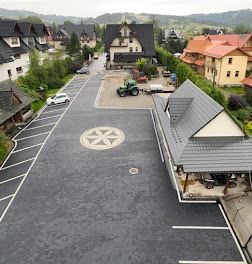 Rozległy plac wyłożony ciemną kostką brukową z dekoracyjnym wzorem krzyża w centralnej części. Widoczne miejsca parkingowe wyznaczone białymi liniami, budynki w tle i sprzęt budowlany. Ujęcie z góry.