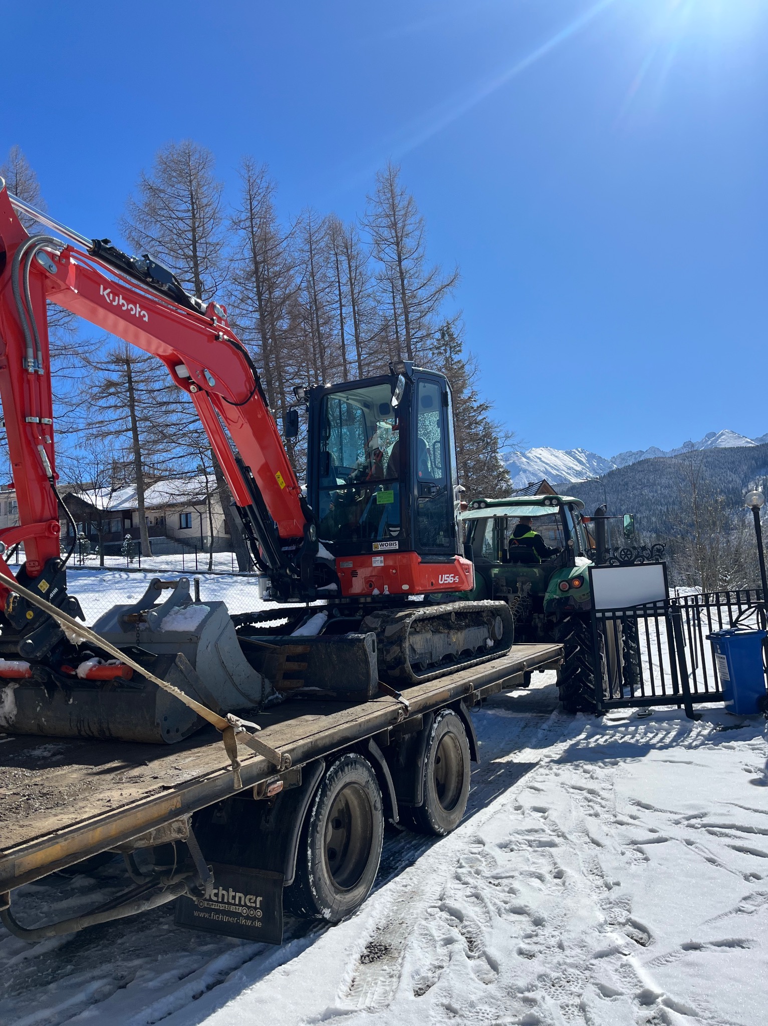 Czerwona mini koparka Kubota U56-5 na lawecie ciężarówki, obok zielony traktor, w tle ośnieżone góry i budynki w zimowej scenerii.