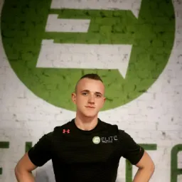 Młody mężczyzna w czarnej koszulce z logo 'Elite Gym&Fitness' na tle białej ceglanej ściany z dużym zielonym logo firmy.