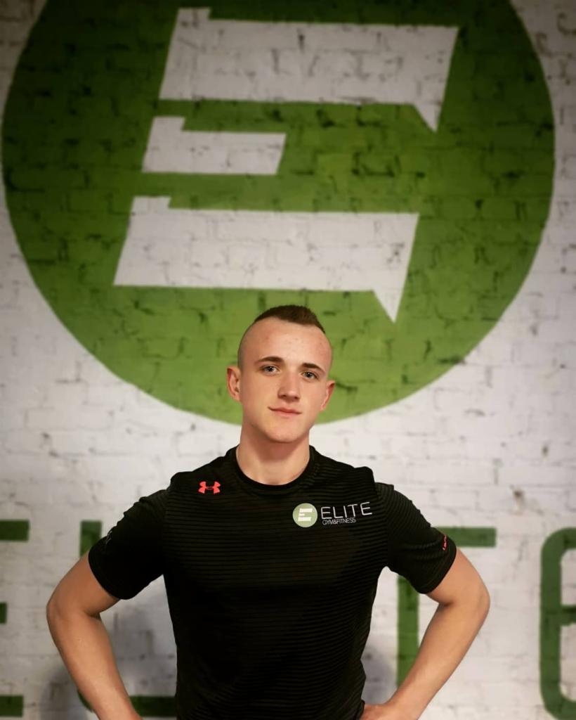 Młody mężczyzna w czarnej koszulce z logo 'Elite Gym&Fitness' na tle białej ceglanej ściany z dużym zielonym logo firmy.