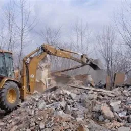 Żółta koparka z wysięgnikiem rozbija fragment ściany budynku na stertę gruzu, w tle drzewa bez liści.