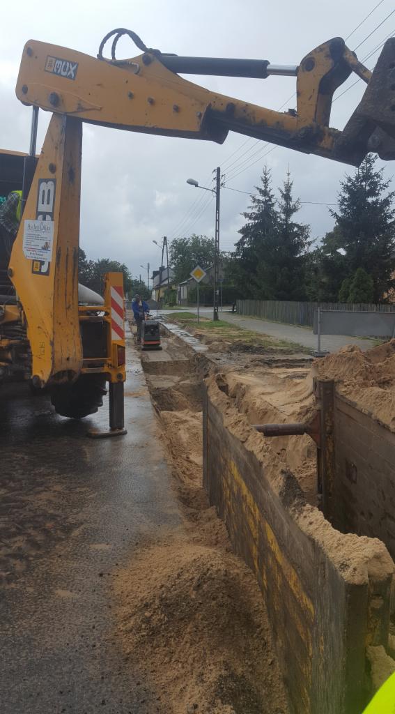Żółta koparko-ładowarka JCB przy wykopie zabezpieczonym szalunkiem, w tle pracownik zagęszcza grunt ubijarką.