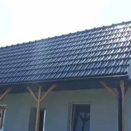 Gotowy dach pokryty ciemną dachówką ceramiczną, z widocznymi drewnianymi elementami konstrukcyjnymi i rynnami w kolorze antracytowym, oświetlony jasnym słońcem.