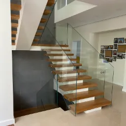 Nowoczesne schody z drewnianymi stopniami i szklaną balustradą w jasnym wnętrzu, widoczne oświetlenie punktowe i galeria zdjęć na ścianie.