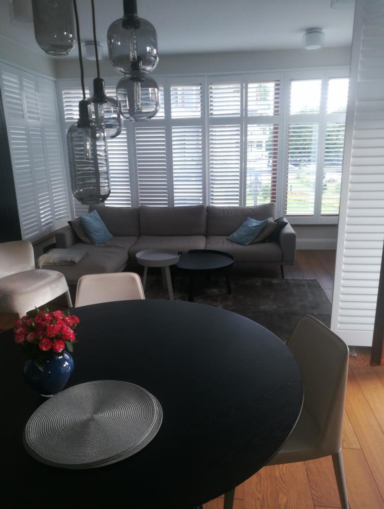 Jasne wnętrze salonu z dużym narożnikiem, okna z białymi żaluzjami, czarny okrągły stół z dekoracją kwiatową, nowoczesne lampy wiszące.