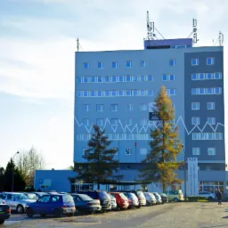 Wielopiętrowy, szary budynek z antenami na dachu, z parkingiem pełnym samochodów przed nim, widziany w słoneczny dzień.