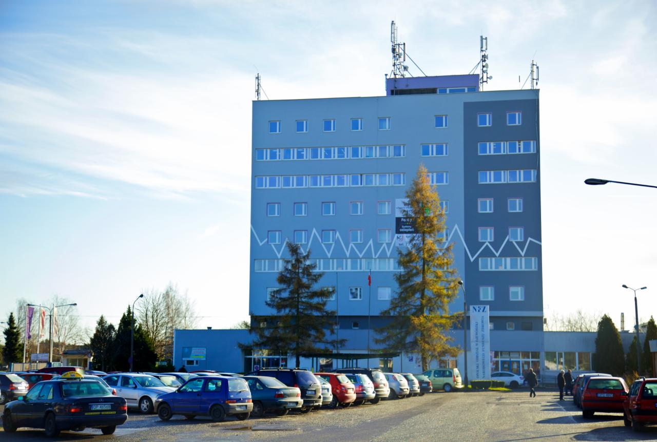 Wielopiętrowy, szary budynek z antenami na dachu, z parkingiem pełnym samochodów przed nim, widziany w słoneczny dzień.