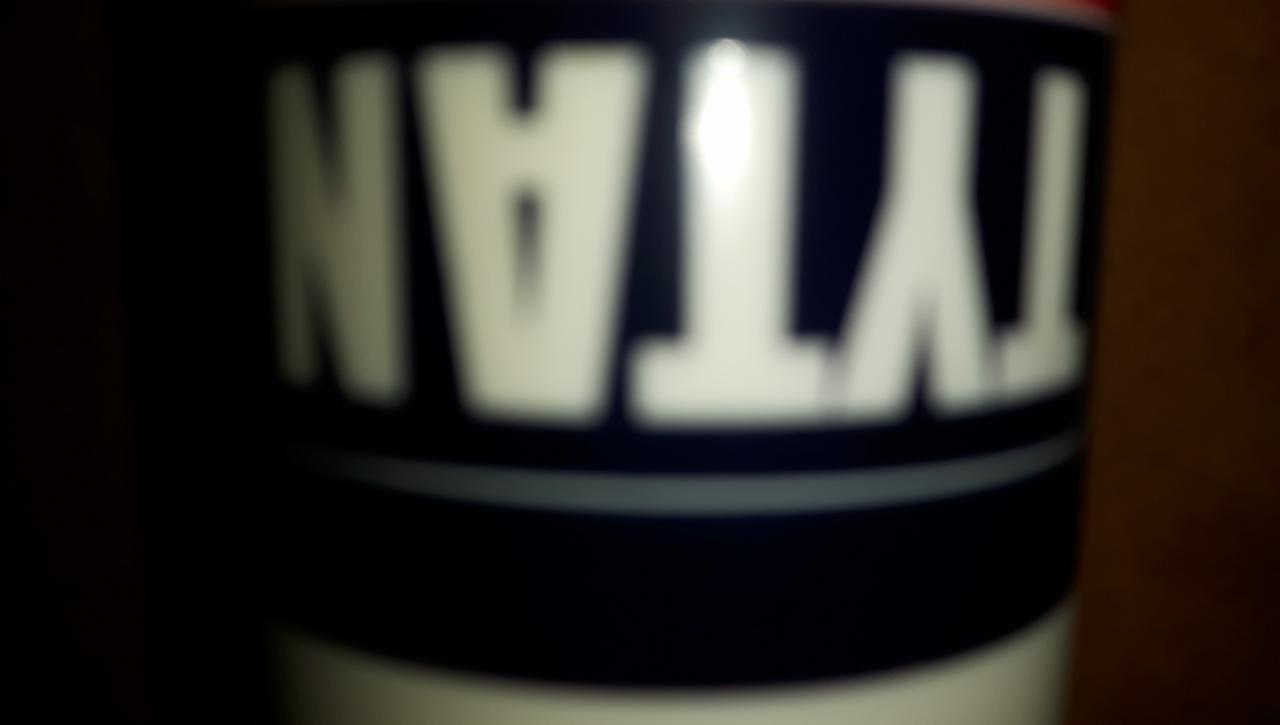 Niejasne ujęcie puszki z pianką montażową Tytan, z widocznym fragmentem etykiety w kolorach biało-granatowo-czerwonym, na tle ciemnego, drewnianego podłoża.