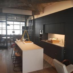 K&K BUD Usługi Remontowo Budowlane Malowanie /Tapetowanie/Zabudowy GK/Docieplanie/Ścianki Działowe itp - Wnętrze kuchni w trakcie remontu: czarne szafki kuchenne, wyspa z szarym blatem, drewniana podłoga, metalowa ścianka działowa z szybami, drabina w tle, widoczne ślady prac remontowych.