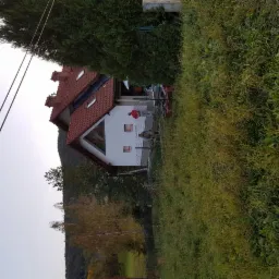 Dom jednorodzinny z białą elewacją i czerwoną dachówką, widoczny podczas prac wykończeniowych na zewnątrz, z elementami rusztowania i osobą w czerwonej kurtce.