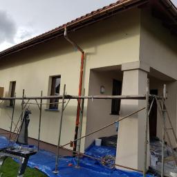 K&K BUD Usługi Remontowo Budowlane Malowanie /Tapetowanie/Zabudowy GK/Docieplanie/Ścianki Działowe itp - Dom jednorodzinny w trakcie ocieplania elewacji, widoczne rusztowania, zabezpieczona folią podstawa budynku, świeżo otynkowana ściana w kolorze beżowym, brązowe rynny i okna.