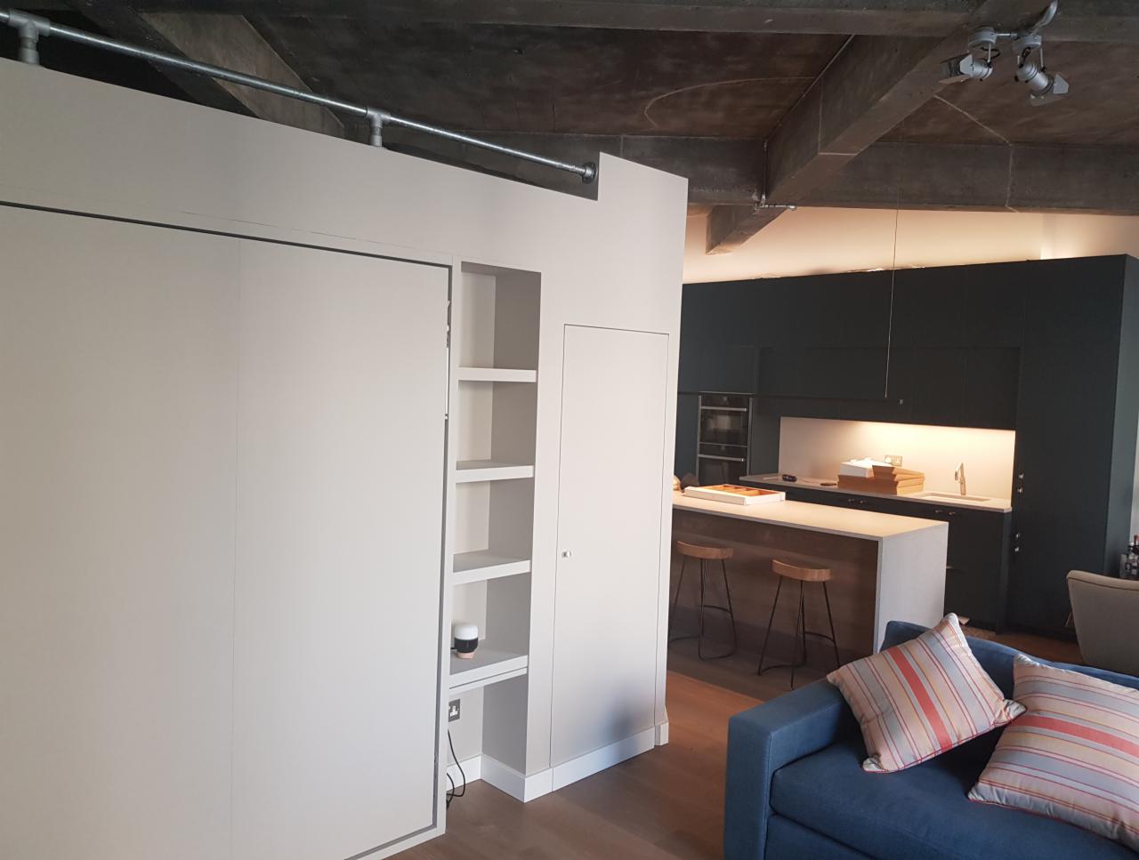 Nowoczesne wnętrze mieszkania z ukrytą szafą w zabudowie, otwartą kuchnią z wyspą i elementami surowego betonu na suficie.