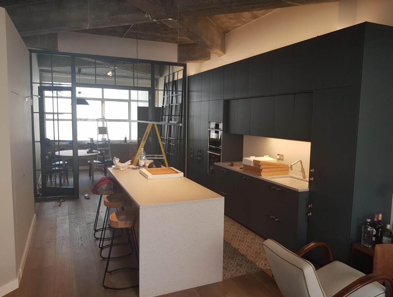 Wnętrze kuchni w trakcie remontu: czarne szafki kuchenne, wyspa z szarym blatem, drewniana podłoga, metalowa ścianka działowa z szybami, drabina w tle, widoczne ślady prac remontowych.