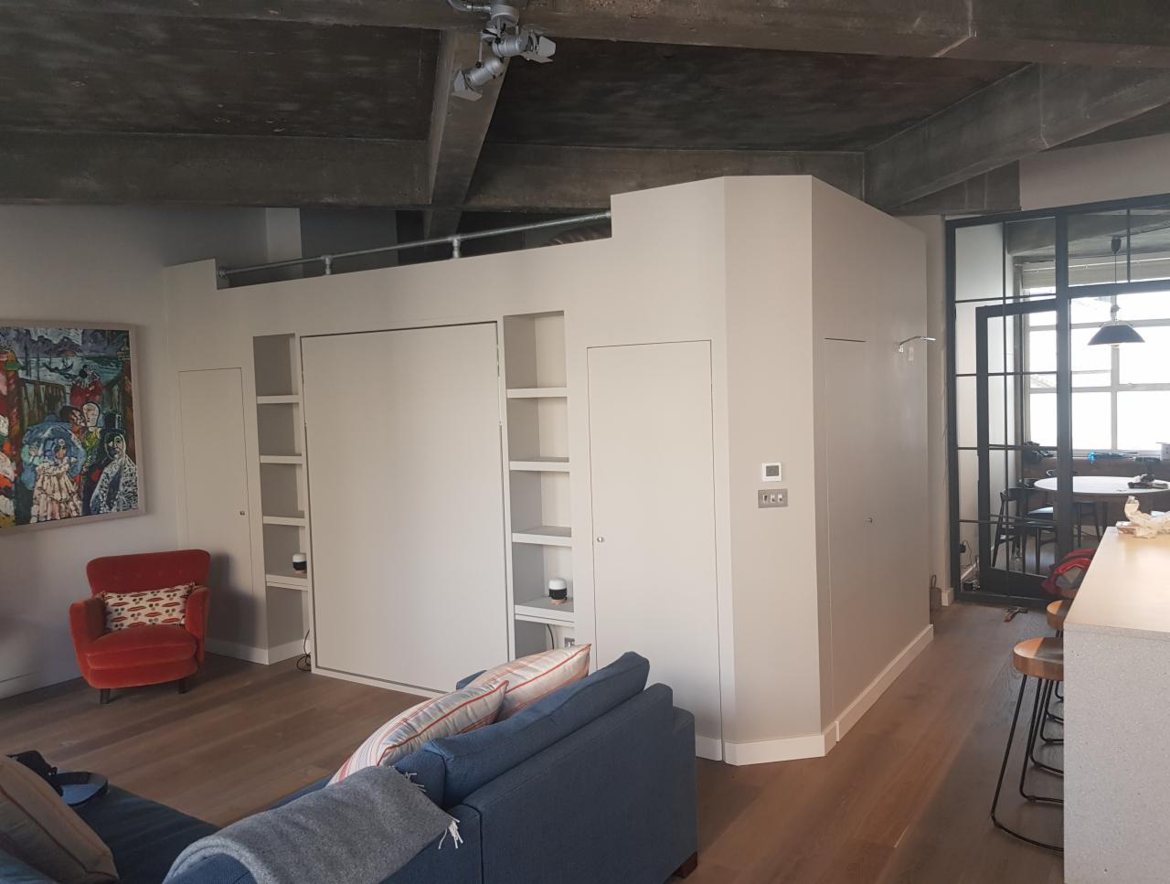 Nowoczesne wnętrze mieszkania z ukrytą szafą wnękową, drewnianą podłogą i betonowym sufitem, widok na przeszkloną ścianę z czarnymi szprosami.