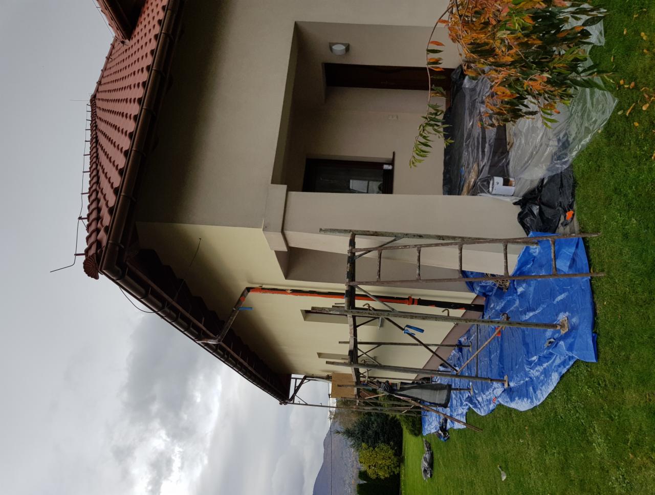 Dom jednorodzinny w trakcie prac elewacyjnych, widoczne rusztowanie, zabezpieczona folią zieleń i fragmenty pomalowanej ściany w odcieniach beżu.