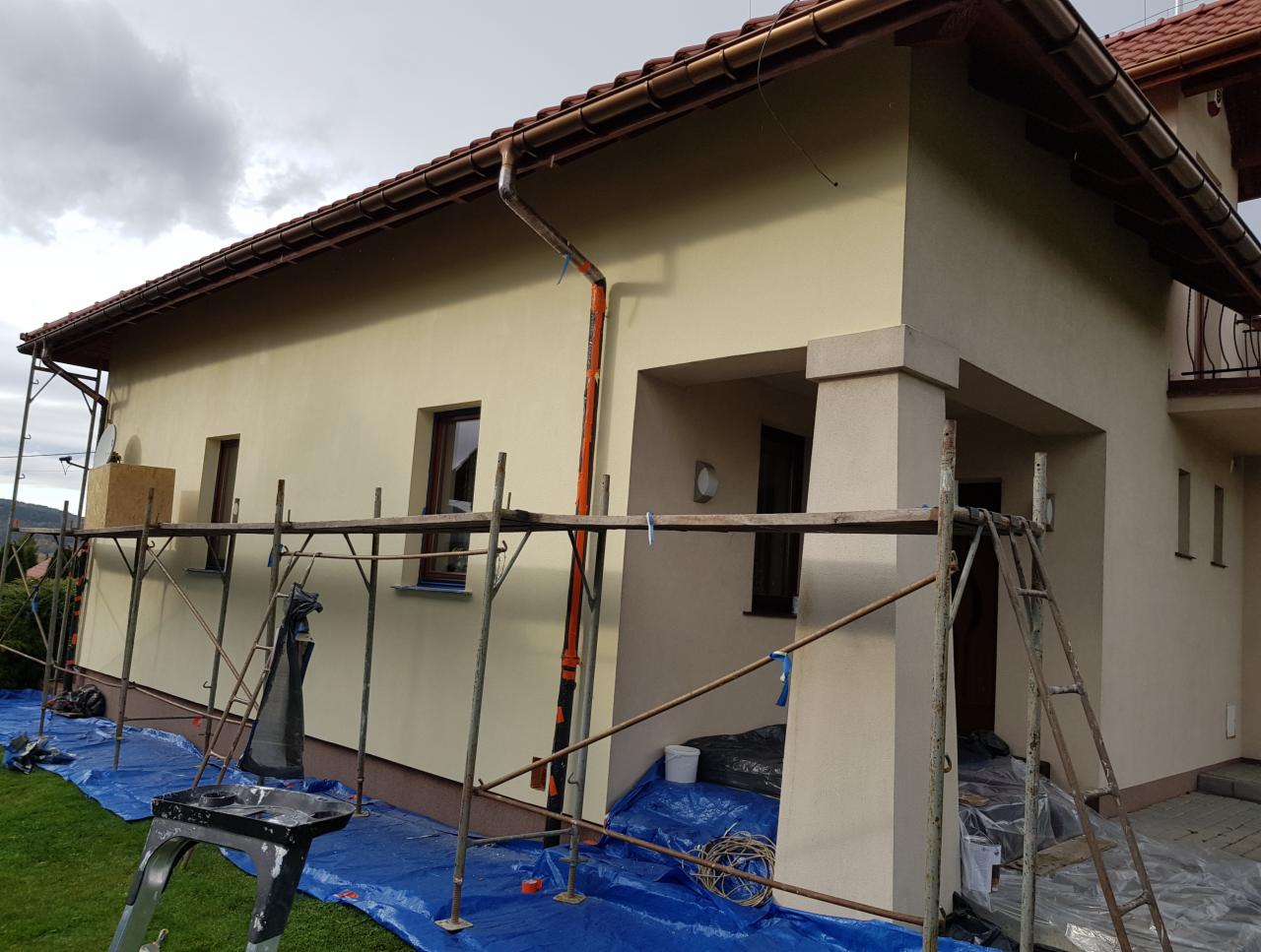 Dom jednorodzinny w trakcie ocieplania elewacji, widoczne rusztowania, zabezpieczona folią podstawa budynku, świeżo otynkowana ściana w kolorze beżowym, brązowe rynny i okna.