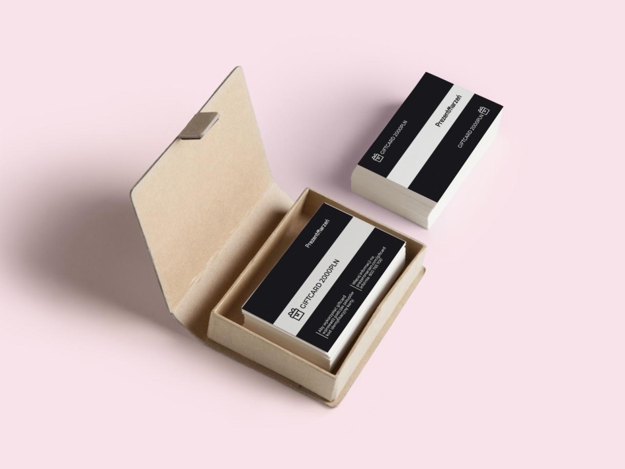 Kartonowe pudełko z kartami podarunkowymi 'Prezentmarzeń' o nominale 2000 PLN, ułożonymi w stos, oraz dodatkowy stos kart obok, na jasnoróżowym tle.