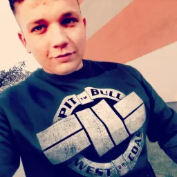 Młody mężczyzna w ciemnej bluzie z dużym logo 'Pit Bull West Coast', pozujący na tle ściany w biało-pomarańczowe pasy. Widoczny fragment drzewa po lewej stronie.
