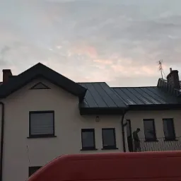 Dom z nowym, ciemnoszarym dachem z blachy na tle pochmurnego nieba. Widoczny fragment czerwonego samochodu na dole kadru i sylwetka osoby na balkonie.