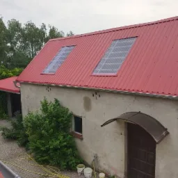 Czerwony dach z blachy trapezowej z dwoma świetlikami na budynku gospodarczym z szarą elewacją i widocznymi rynnami