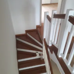 Klatka schodowa z drewnianymi schodami w kolorze ciemnego brązu z białymi podstopnicami i białą balustradą, widok z góry, na jednym ze stopni leży rolka papieru i butelka.