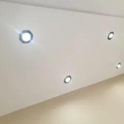 Biały sufit pod skosem z czterema punktowymi lampami LED o ośmiokątnych obudowach, oświetlającymi beżową ścianę. Minimalizm i nowoczesne wykończenie.