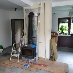 Instalacja kominka z czarnym wkładem i rurą dymową, zabezpieczona folią aluminiową, poziomica oparta o ścianę, drabina i narzędzia obok, kartony na podłodze.