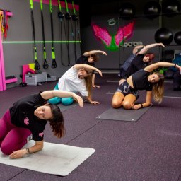 Fit & Fizjo Team - trening personalny, fizjoterapia, masaże Gdańsk - Grupa osób, w tym jeden mężczyzna, wykonuje ćwiczenia rozciągające na matach w jasnym, nowoczesnym fitness studio z neonowymi akcentami i sprzętem do ćwiczeń w tle.