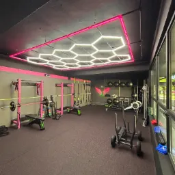 Nowoczesna siłownia z czarno-różowym wystrojem, wyposażona w sprzęt do ćwiczeń siłowych i cardio, oświetlona geometryczną lampą sufitową z różowym obramowaniem.